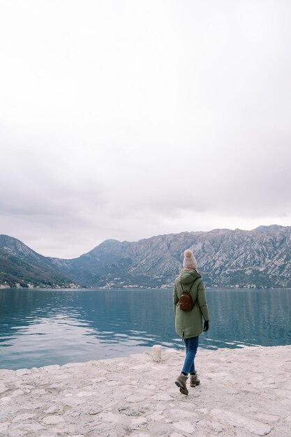 Une femme avec un sac à dos se promène le long de la jetée près de la mer et regarde les montagnes à l'arrière