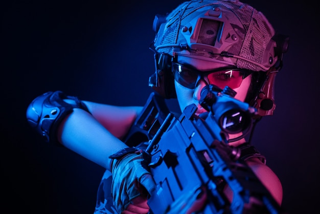 Femme en salopette militaire airsoft posant avec une arme à feu dans ses mains sur un mur sombre dans la brume en néon