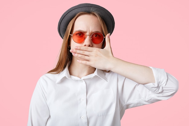 Une Femme Sans Expression Et En Colère Couvre La Bouche Alors Qu'elle Tente De Garder Le Silence, Porte Un Chapeau élégant Et Des Nuances élégantes, Isolé Sur Le Mur Rose Du Studio