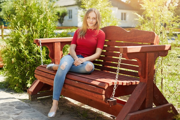 Femme sans soucis avec des cheveux raides portant un pull rouge et un jean à la mode assis à un banc en bois confortable dans le parc en profitant du soleil