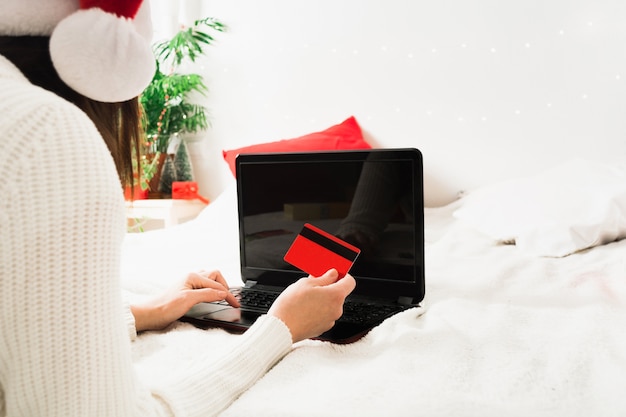 Femme sans visage en bonnet de Noel allongé sur le lit à la maison avec ordinateur portable et carte en plastique