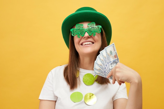 Une femme satisfaite et joyeuse portant un chapeau de leprechaun vert et des lunettes de trèfle posant isolée sur un fond jaune tenant une grosse somme d'argent appréciant sa récompense