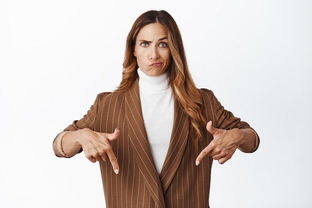 Femme sceptique PDG de l'entreprise grimaçant et regardant avec incrédulité pointant du doigt la publicité ci-dessous debout en costume marron sur fond blanc