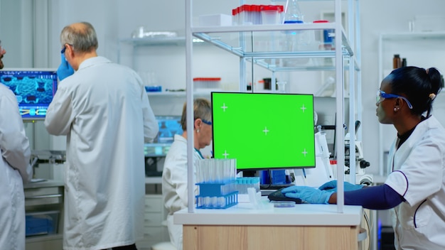 Femme scientifique africaine tapant sur ordinateur avec une maquette verte dans un laboratoire moderne équipé. Équipe multiethnique de microbiologistes effectuant des recherches sur les vaccins écrivant sur un appareil avec clé chroma, affichage isolé.