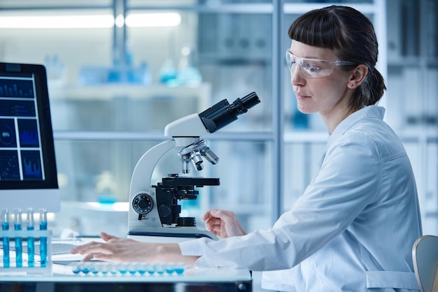 Femme scientifique assise à table en laboratoire et examinant des échantillons dans des éprouvettes au microscope