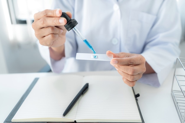 Femme scientifique tenant un compte-gouttes pour déposer un échantillon de solution sur la plaque de verre pour microscope et liquide bleu dans un tube à essai tout en travaillant à l'analyse et au développement d'un vaccin contre le coronavirus en laboratoire