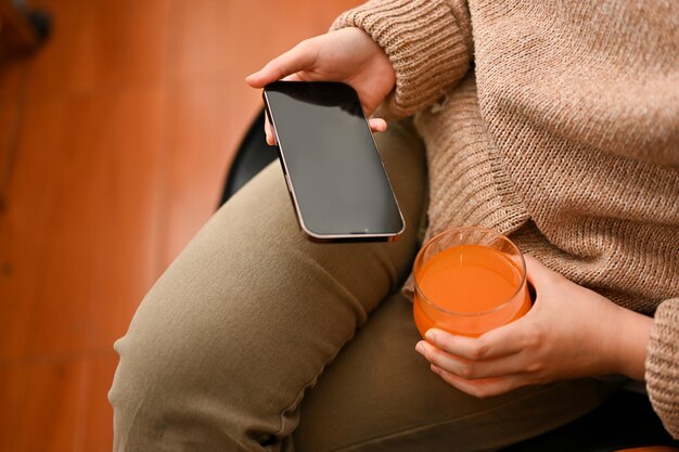 Une femme se détend assise sur un fauteuil en sirotant du jus d'orange et en utilisant un téléphone portable