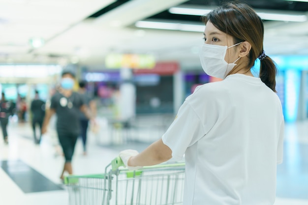 La femme se protège contre l'infection avec le masque chirurgical et les gants, avec panier d'achat pour faire les courses au supermarché après la pandémie de coronavirus.