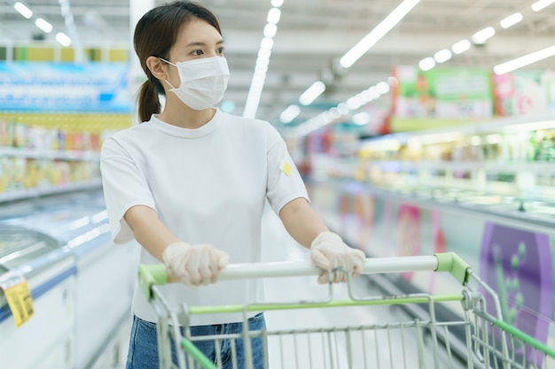 La femme se protège contre l'infection avec le masque chirurgical et les gants, avec panier d'achat pour faire les courses au supermarché après la pandémie de coronavirus.