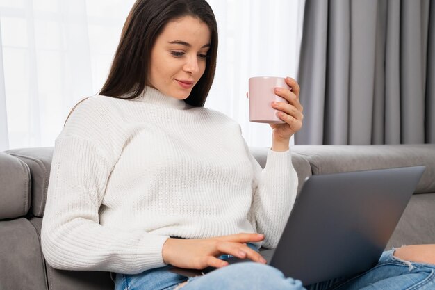 Femme se relaxant avec une tasse de thé à la maison à l'aide d'un ordinateur portable dans un appartement moderne