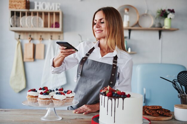 Une femme se tient dans la cuisine et prend une photo de ses biscuits et de sa tarte maison à l'aide d'un téléphone.