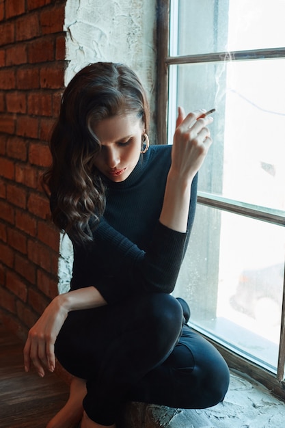 La femme se tient à la fenêtre et fume une cigarette. Fille avec beau maquillage, cosmétiques naturels