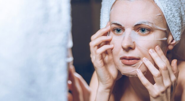 Femme se tient près d'un miroir avec une serviette sur la tête et met un masque cosmétique