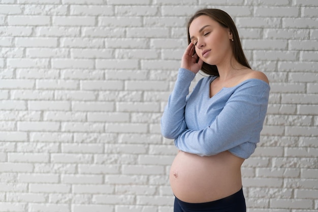 Une femme se tient près d'un mur de briques blanches Tient sa tête avec sa main Concept d'expérience de grossesse