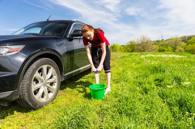 Femme avec un seau vert essorant une éponge savonneuse et lavant un véhicule de luxe noir dans un champ vert par une belle journée ensoleillée avec un ciel bleu