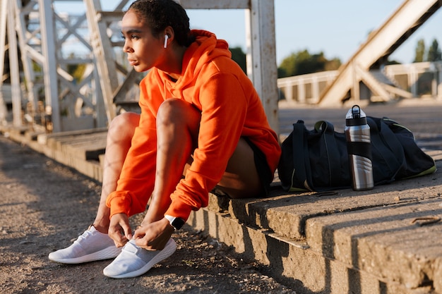 femme séduisante afro-américaine en vêtements de sport utilisant des oreillettes et attachant des lacets assis sur le vieux pont