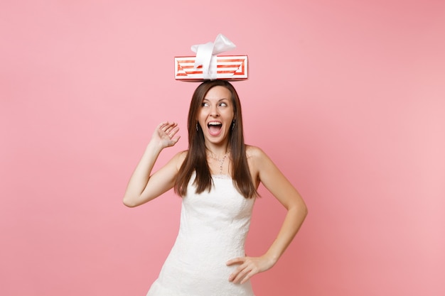 Femme séduisante drôle en robe blanche debout, tenant une boîte rouge avec un cadeau, présente sur la tête