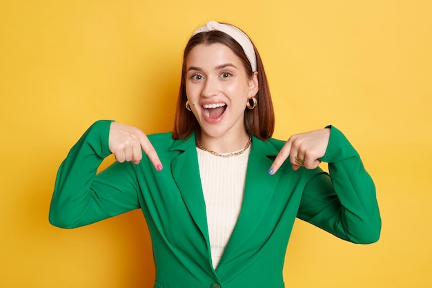 Femme séduisante ravie portant une veste verte posant isolée sur fond jaune pointant vers le haut demandant de s'abonner à son blog regardant la caméra avec un visage heureux