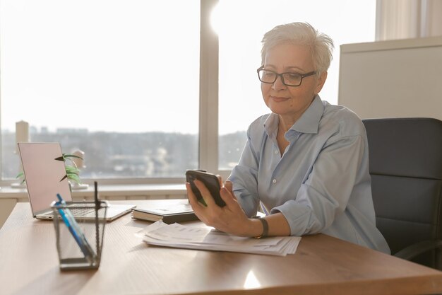 Femme senior d'âge moyen européenne élégante et confiante utilisant un smartphone sur le lieu de travail élégant matu plus âgé