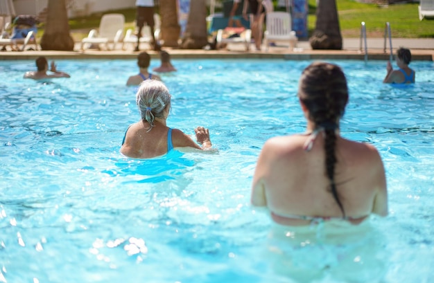 Femme senior âgée aux cheveux gris, portant un maillot de bain bleu faisant de l'aquagym dans la piscine de l'hôtel, vue de derrière