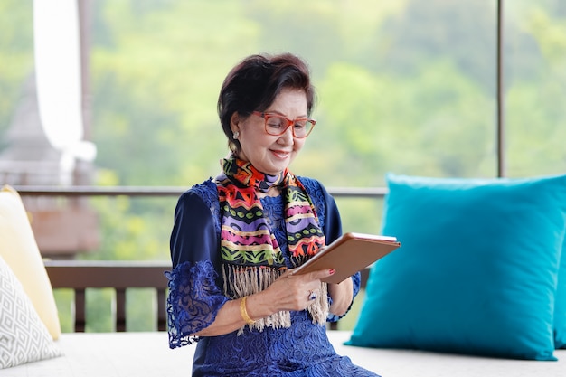 Femme senior asiatique assise sur le canapé et à l'aide d'une tablette