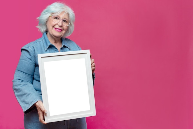 Femme senior aux cheveux gris tenant un cadre photo vide sur fond rose