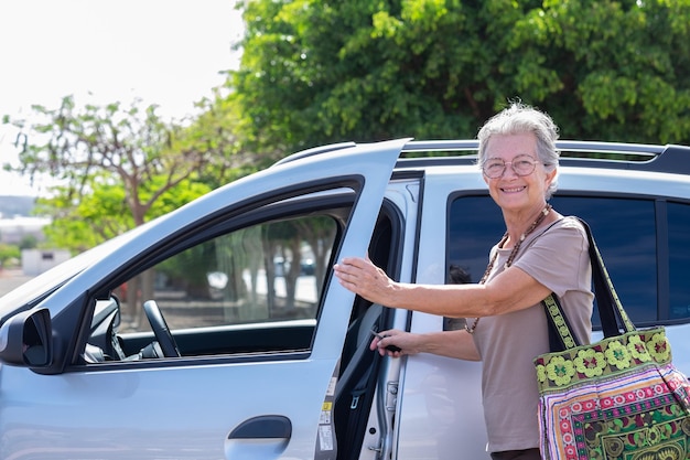 Une femme senior caucasienne souriante entrant dans sa voiture une grand-mère heureuse avec un grand sac fait main ouvre la porte pour entrer dans le véhicule prêt à conduire