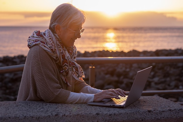 Femme senior caucasienne souriante utilisant un ordinateur portable dans le travail à distance à la plage Coucher de soleil doré sur fond