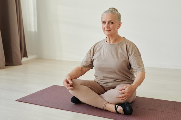 Femme senior contemporaine en vêtements de sport assise en position du lotus sur un tapis tout en pratiquant des exercices de yoga relaxants à loisir