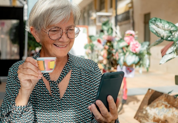 Femme senior détendue assise au café en plein air à l'aide de son smartphone tout en dégustant une tasse de café expresso