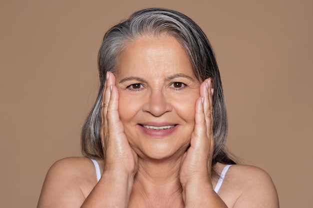 Une femme senior européenne joyeuse aux cheveux gris touche le visage avec les mains et bénéficie du résultat de la procédure de spa