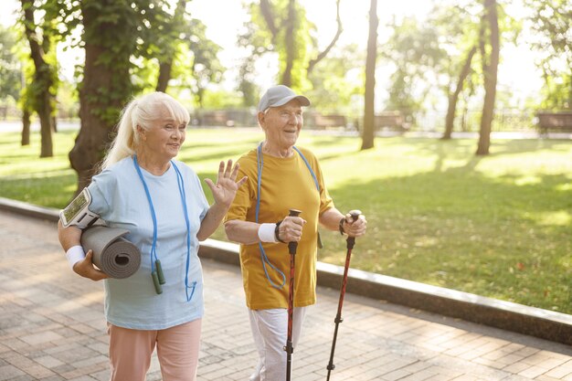 Femme senior positive avec tapis et homme avec des bâtons pour la marche nordique dans un parc verdoyant