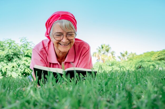 Femme senior souriante en rose allongée sur l'herbe dans un parc public ayant des moments de détente en lisant un livre