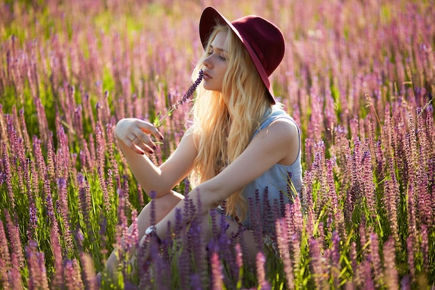 Femme sensuelle aux cheveux blonds portant un chapeau posant dans un champ floral derrière un beau fond de coucher de soleil