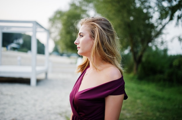 Femme sensuelle blonde en robe rouge marsala posant sur la plage de la station balnéaire.