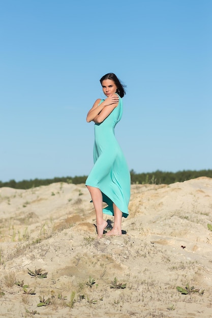 Femme sensuelle mince au corps entier avec des cheveux volants portant une longue robe turquoise en se tenant debout dans le vent sur la côte