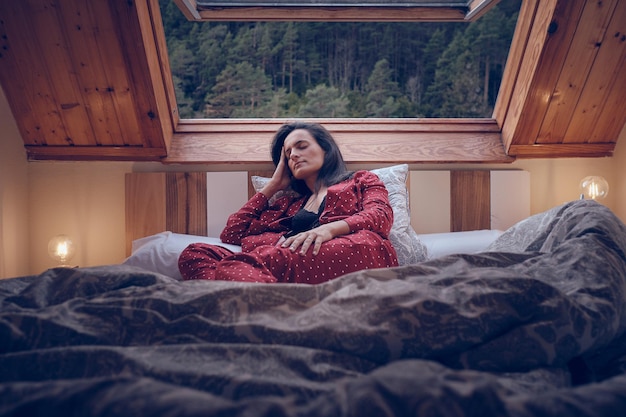 Femme sereine en pyjama allongée sur un lit confortable près de la fenêtre et dormant tout en passant du temps dans la maison des hautes terres