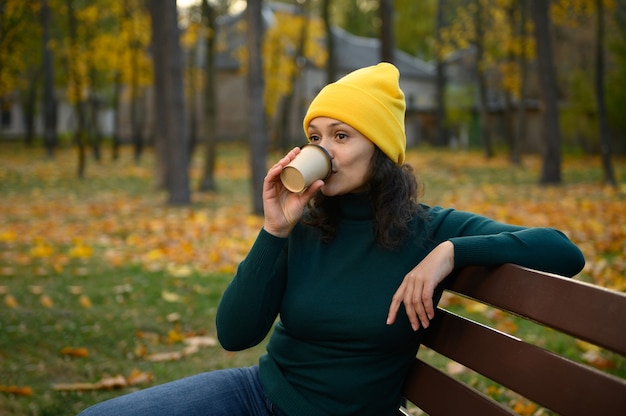 Femme sereine et ravie dans un chapeau tricoté jaune en laine chaude, assise sur un banc de parc et buvant du café dans une tasse de papier artisanal à emporter, se reposant dans un parc forestier automnal avec des érables en arrière-plan