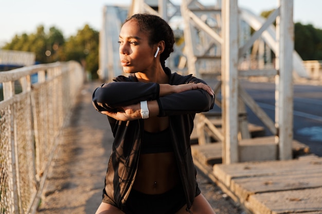 femme sérieuse afro-américaine en tenue de sport utilisant des écouteurs et faisant des exercices tout en s'entraînant sur le vieux pont