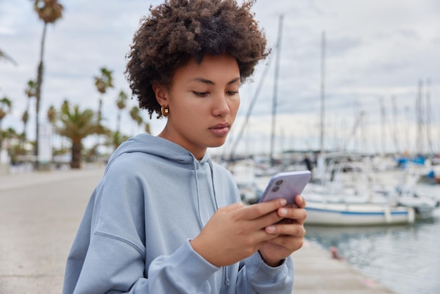Une femme sérieuse aux cheveux bouclés porte un sweat-shirt bleu utilise un téléphone portable surfe sur les médias sociaux vérifie les notifications pose contre le port de la mer vêtu d'un sweat à capuche décontracté Les gens et le concept de style de vie moderne