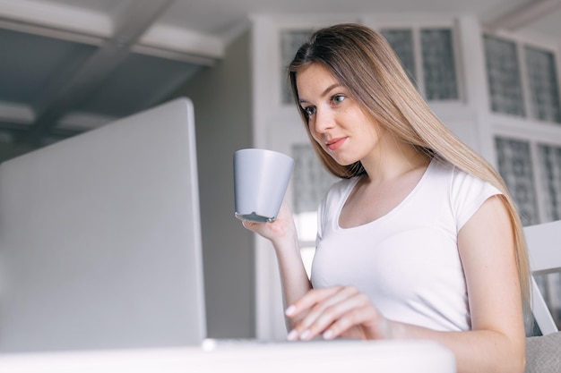 Une femme sérieuse boit du café à l'aide d'un ordinateur dans la cuisine à la maison w