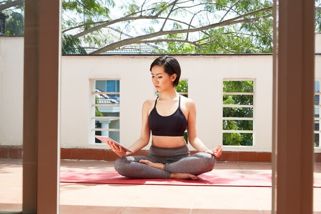 Femme sérieuse et calme assise sur un tapis de yoga en position du lotus et regardant une vidéo n méditation sur un ordinateur tablette sur son balcon