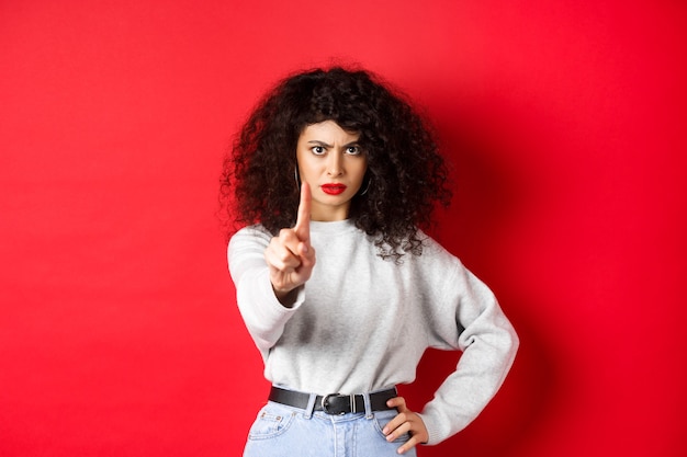 Femme sérieuse et confiante dire non, tendre un doigt pour vous arrêter, interdire quelque chose de mal, debout déterminée sur fond rouge.