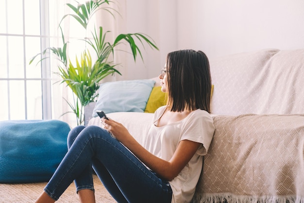 Femme sérieuse dans des lunettes de messagerie texte à l'aide d'un téléphone portable alors qu'elle était assise par terre dans le salon de sa maison. Belle dame passe son temps libre à l'aide d'un smartphone à la maison