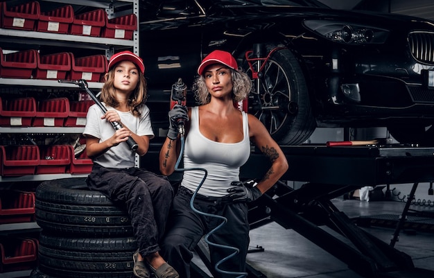 Une femme sérieuse expérimentée et sa petite aide posent pour le photographe au service automobile sombre en tant qu'équipe formidable.