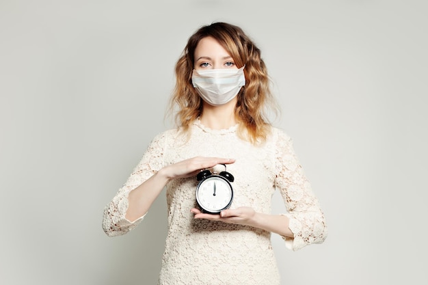 Une femme sérieuse avec un masque médical protecteur et un réveil blanc.