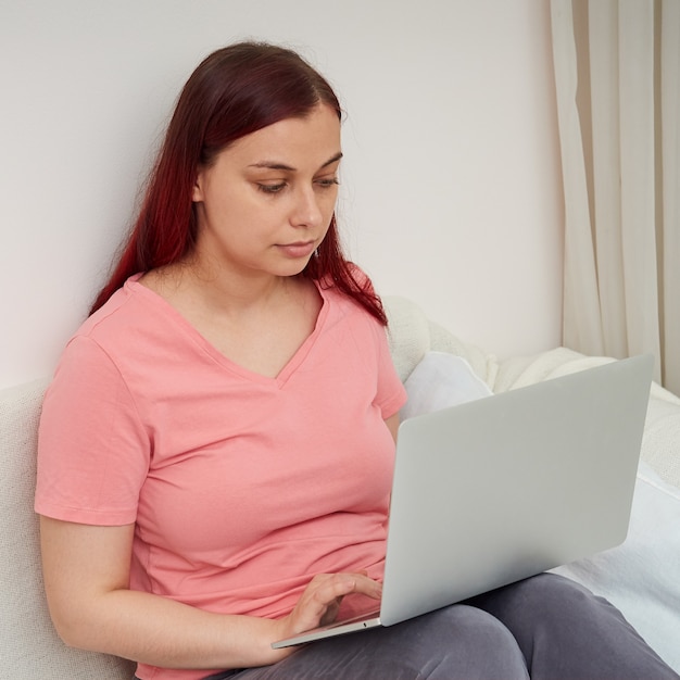 Femme sérieuse en pyjama assise sur le lit travaille à distance avec un ordinateur portable fait des achats sur internet...
