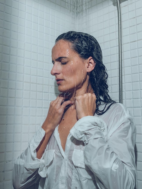 Femme sérieuse et réfléchie en chemise blanche et avec des cheveux noirs mouillés, debout, les yeux fermés, sous l'eau qui coule dans la cabine de douche