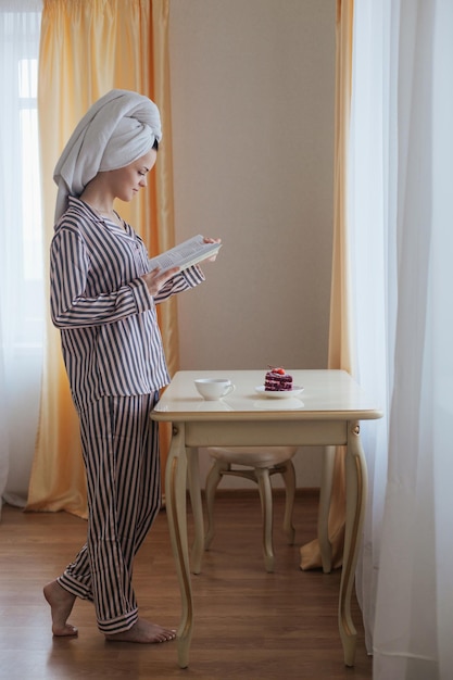 Femme en serviette se tient près de la table lire un livre intéressant boire du thé avec un gâteau le matin