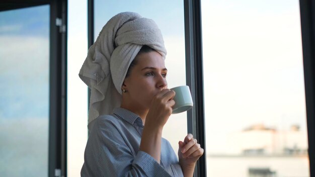 Femme avec une serviette sur la tête buvant du café en arrière-plan du concept de fenêtre belle jeune femme